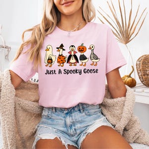 May include: A light pink t-shirt featuring a graphic of five cartoon geese dressed in Halloween costumes. The geese are wearing a ghost costume, witch costume, vampire costume, pumpkin costume, and skeleton costume. The text "Just A Spooky Goose" is below the geese.