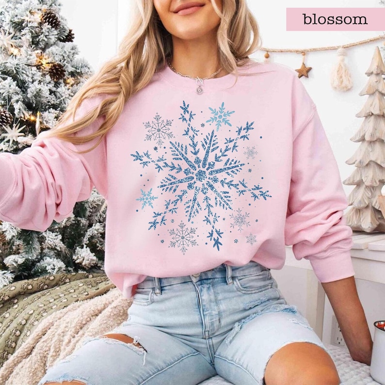 May include: A light pink sweatshirt featuring a large blue snowflake design. The snowflake has a glittery appearance and is surrounded by smaller snowflakes. The word "blossom" is printed in a pink rectangle in the upper right corner.