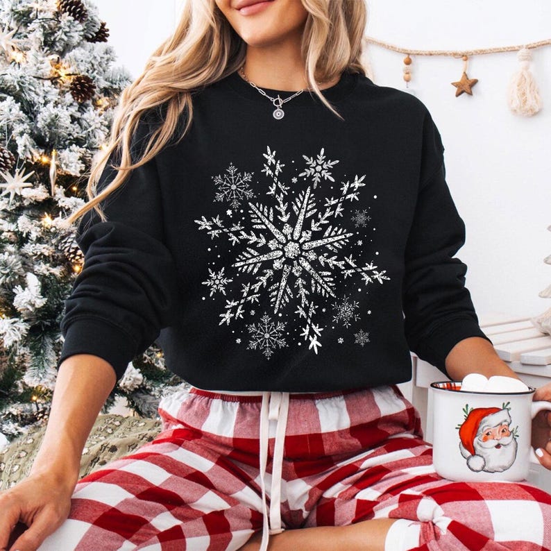 May include: Black sweatshirt featuring a large, detailed white snowflake design. The snowflake is the focal point, surrounded by smaller snowflakes. The person is wearing red and white plaid pajama pants. A mug with a Santa Claus illustration is held in the hand.