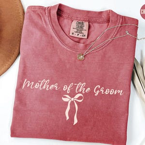 May include: Crimson red t-shirt with the text "Mother of the Groom" and a pink bow.