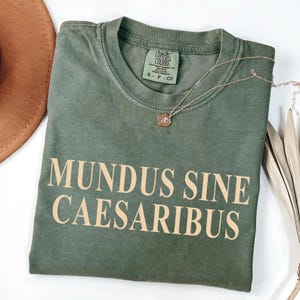 May include: A green t-shirt with the text "MUNDUS SINE CAESARIBUS" in a light beige color. The shirt is made of a soft, comfortable material and has a relaxed fit.