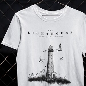 Könnte beinhalten: Weißes T-Shirt mit einer Schwarzweißillustration eines Leuchtturms mit Vögeln, die um ihn herum fliegen. Der Text "THE LIGHTHOUSE" ist über dem Bild gedruckt. Unter dem Bild steht "From Robert Eggers, Director of The VVitch."