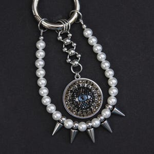 May include: A keyring featuring a large silver ring attached to two strands of white faux pearls and a central silver chain. The chain holds a circular pendant with a blue eye graphic surrounded by black and gold crystals and four silver spikes.