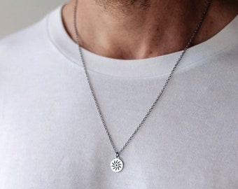 Könnte beinhalten: Eine silberfarbene Metallhalskette mit einem kleinen, runden Anhänger, der ein ausgeschnittenes Sonnenmotiv zeigt. Der Anhänger hängt an einer zarten, passenden silberfarbenen Kette auf einem schlichten weißen Stoff.