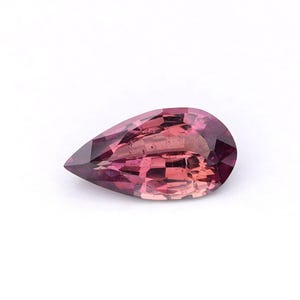 May include: A pear-shaped gemstone with a gradient of colors, from deep burgundy to light pink. The gem has a faceted surface, catching light and displaying a range of hues. The stone is set against a white background, highlighting its color and shape.