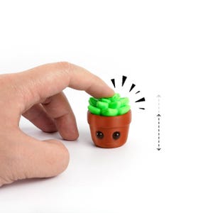 Puede incluir: Una pequeña maceta marrón de estilo caricatura con una planta suculenta verde encima. La maceta tiene dos ojos pequeños y redondos y una boca sonriente. Un dedo toca la planta, con líneas radiantes que indican movimiento.