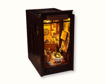 May include: A miniature wooden model of a detective agency storefront with a lit interior. The storefront has a sign that reads "Detective Agency". The interior is furnished with a desk, chairs, and a bookshelf.