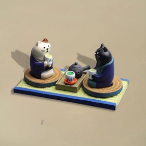 Könnte beinhalten: Dekorative Figur mit zwei Katzen in traditioneller japanischer Kleidung, die an einer Teezeremonie teilnehmen. Die Katzen, eine weiße und eine schwarze, halten Teetassen. Eine kleine Teekanne und Tassen stehen auf einem Tablett. Die Basis ist eine rechteckige Matte mit blauem Rand.