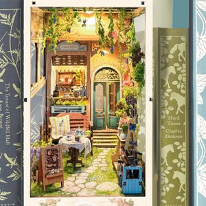 May include: A miniature dollhouse scene with a small cafe, a patio with a table and chairs, and a doorway leading to a garden. The cafe has a sign that says "Flower Cafe".
