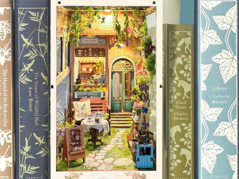 May include: A miniature dollhouse scene with a small cafe, a patio with a table and chairs, and a doorway leading to a garden. The cafe has a sign that says "Flower Cafe".