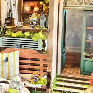 May include: A miniature dollhouse scene with a wooden bench, a planter with green plants and flowers, and a green door with a window. The scene is decorated with butterflies and a small bouquet of flowers.