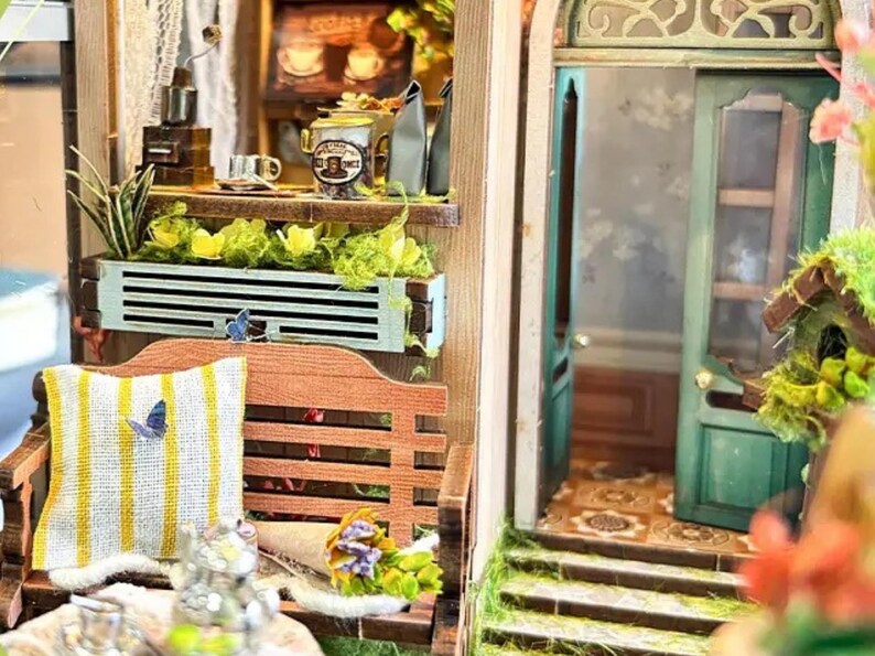 May include: A miniature dollhouse scene with a wooden bench, a planter with green plants and flowers, and a green door with a window. The scene is decorated with butterflies and a small bouquet of flowers.