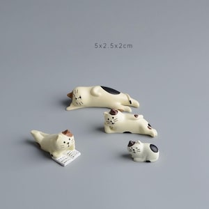 May include: Four cream-colored cat figurines with black and brown markings in various poses. One cat is reading a book, while others are lounging. The text "5x2.5x2cm" is visible. These figurines are ideal for cat lovers.