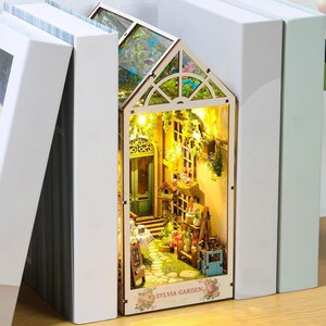 May include: A miniature dollhouse with a glass roof and a light inside. The dollhouse is decorated with flowers and greenery. The dollhouse is labeled "Sylvia Garden".
