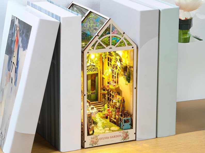 May include: A miniature dollhouse with a glass roof and a light inside. The dollhouse is decorated with flowers and greenery. The dollhouse is labeled "Sylvia Garden".