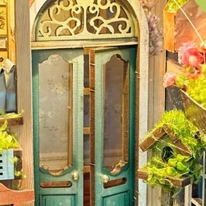 May include: A miniature dollhouse with a green door with glass panels and a decorative wooden frame. The door is open and there are shelves visible inside. There are plants and flowers in pots on the shelves. There is a birdhouse with a green roof and a small bird inside. The dollhouse is made of wood and has a rustic look.