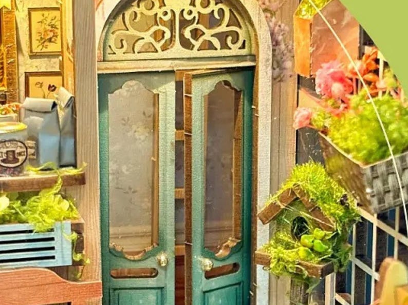 May include: A miniature dollhouse with a green door with glass panels and a decorative wooden frame. The door is open and there are shelves visible inside. There are plants and flowers in pots on the shelves. There is a birdhouse with a green roof and a small bird inside. The dollhouse is made of wood and has a rustic look.