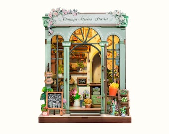 Puede incluir: Una tienda de casa de muñecas en miniatura con una fachada verde y un letrero que dice "Champs-Élysées Florist". La tienda tiene un tema de floristería con arreglos florales y una variedad de plantas exhibidas dentro y fuera de la tienda.