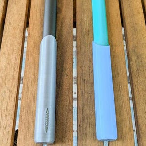 Puede incluir: Dos portalápices impresos en 3D, uno gris y negro, el otro azul claro y turquesa. El soporte gris tiene la palabra "omo" impresa. Los portalápices están sobre una superficie de madera.