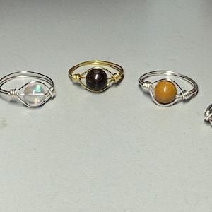 May include: Five wire-wrapped rings with different colored gemstone beads. The rings have silver and gold-colored bands. The gemstones are blue, clear, brown, yellow, and red.