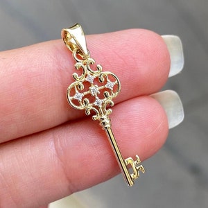 May include: A gold key pendant with a detailed ornate design and small clear stones. The key is on a hand.