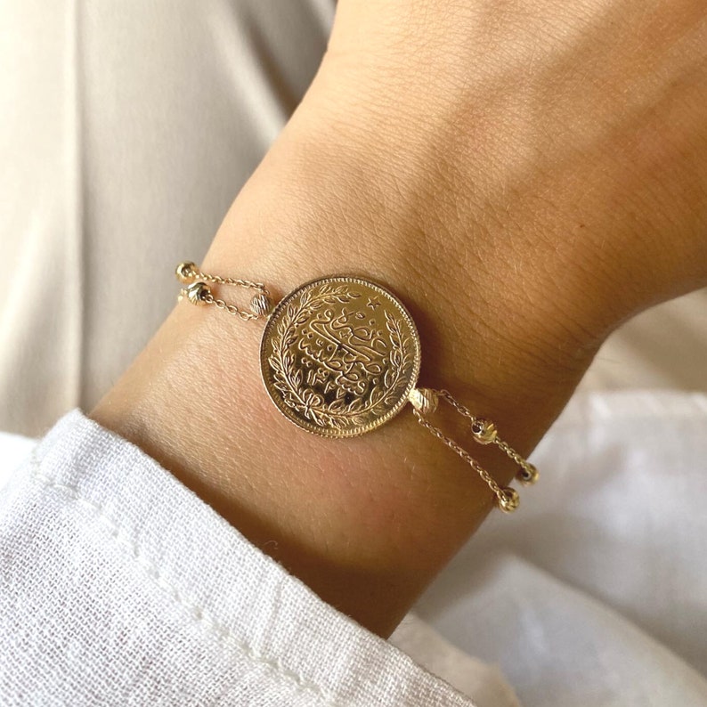 Puede incluir: Un brazalete de oro con una moneda detallada en el centro. La moneda est&aacute; adornada con intrincados grabados y est&aacute; conectada a delicadas cadenas de cuentas. El brazalete es de color oro c&aacute;lido y se lleva en la mu&ntilde;eca.