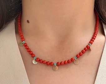 May include: A red beaded necklace with small gold-colored circular charms and turquoise accents. The necklace is worn around the neck and is a close-up shot.