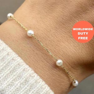 Puede incluir: Una delicada pulsera de cadena dorada con cinco perlas blancas. La pulsera se muestra en una muñeca, con un suéter color crema en el fondo. La imagen incluye una pegatina redonda naranja con las palabras "WORLDWIDE DUTY FREE".