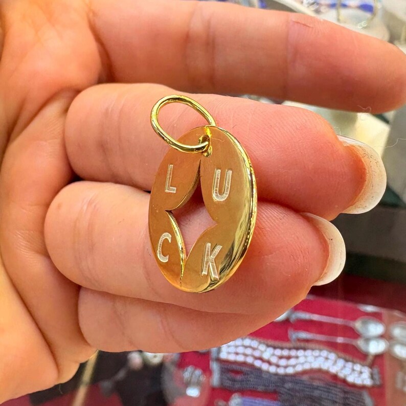 May include: A gold-colored oval pendant with the word "LUCK" cut out in a star shape. The pendant has a small loop at the top for a chain. The background is blurred, but appears to be a display case.