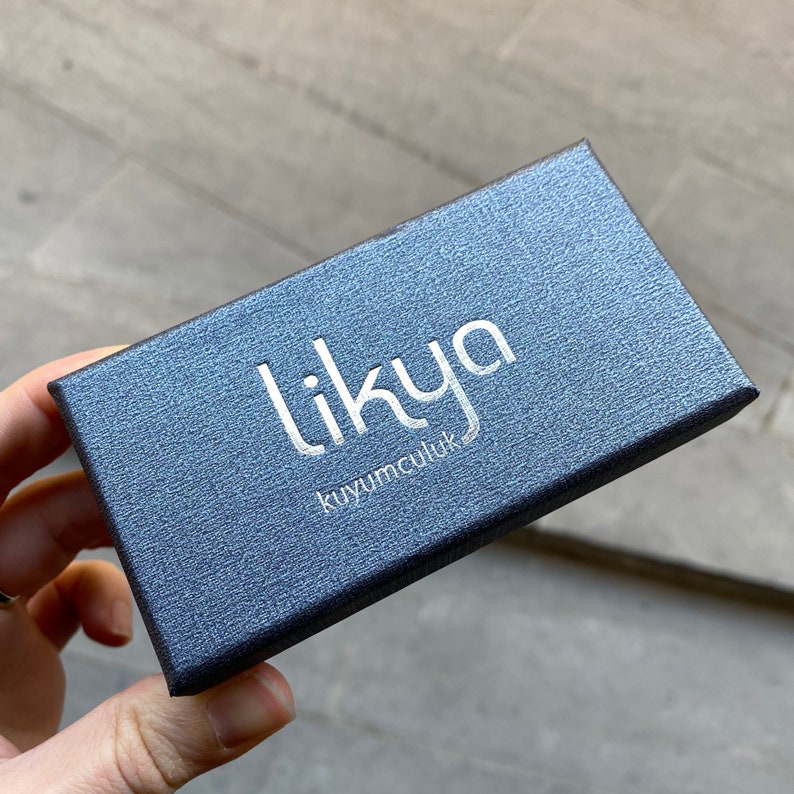 K&ouml;nnte beinhalten: Eine dunkelgraue Schmuckschatulle mit dem Text "likyya kuyumculuk" in Silber auf der Oberseite.
