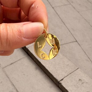 May include: A gold-colored circular pendant with the word "LUCK" inscribed. The pendant has a diamond-shaped cutout in the center. The letters are in a simple, sans-serif font. The pendant is held up by a hand.