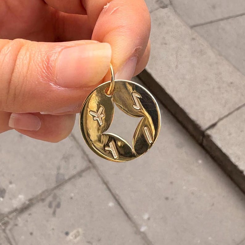 May include: A gold-colored pendant with a circular shape and a star-shaped cutout in the center. The pendant features stylized symbols around the cutout. The pendant is suspended from a small loop.