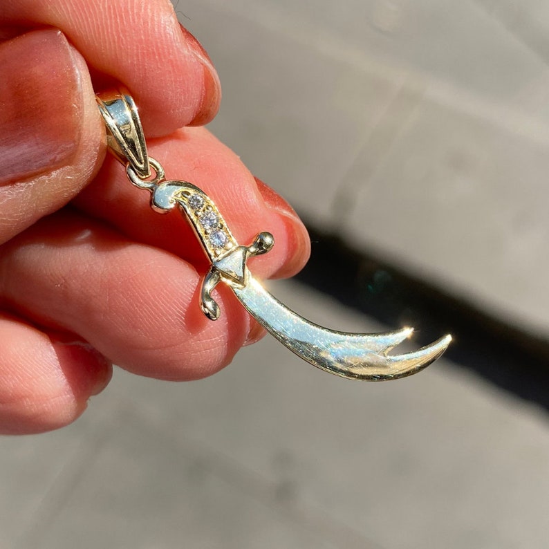 K&ouml;nnte beinhalten: Ein goldfarbener Anh&auml;nger in Form eines gebogenen Schwertes mit einer kleinen Schlaufe oben f&uuml;r eine Kette. Das Schwert hat einen dekorativen Griff mit drei kleinen, klaren Edelsteinen. Die Klinge hat ein gebogenes Design.