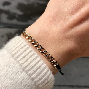 Puede incluir: Una pulsera de cadena dorada con un diseño de eslabones de bordillo. La pulsera se lleva en la muñeca y el cierre es un cordón negro. La muñeca está cubierta por un suéter blanco.