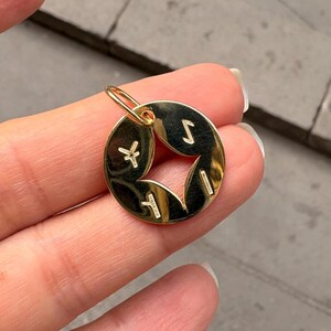May include: A gold-colored pendant with a circular shape and a star-shaped cutout in the center. The pendant has four symbols around the star. The pendant is suspended from a small gold loop.