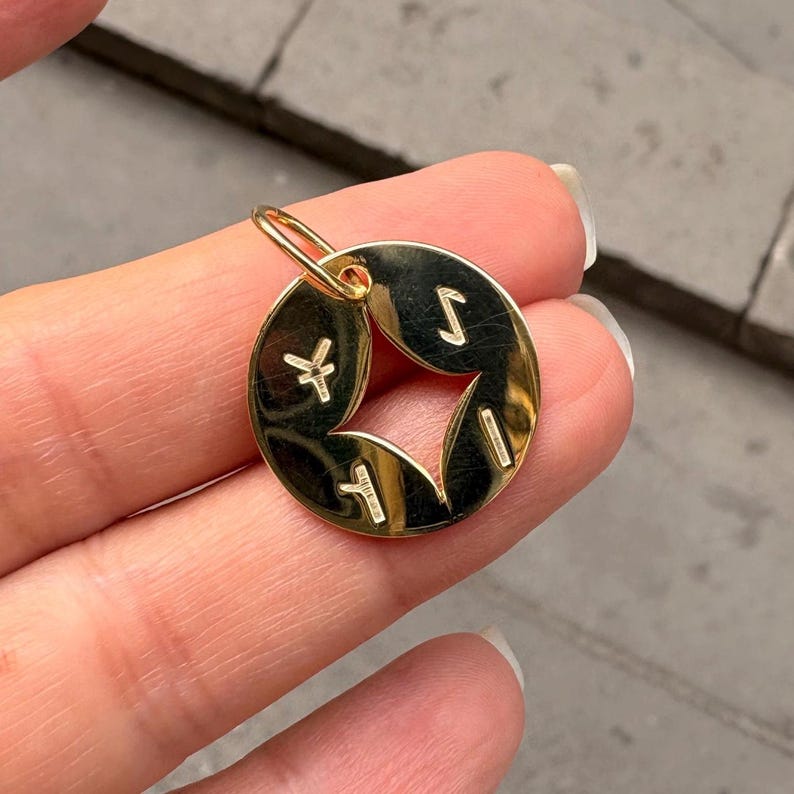 May include: A gold-colored pendant with a circular shape and a star-shaped cutout in the center. The pendant has four symbols around the star. The pendant is suspended from a small gold loop.