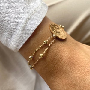 Puede incluir: Una pulsera de monedas de oro con cadenas de cuentas colgantes. La moneda tiene un dise&ntilde;o detallado. La pulsera se usa en una mu&ntilde;eca, con una manga blanca en el fondo. La pulsera est&aacute; hecha de metal color oro.