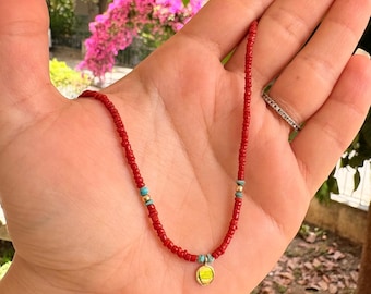 May include: A red beaded necklace with turquoise and gold accents, and a small, round, yellow pendant. The necklace is held in a hand, with a ring on the finger. The background features pink flowers and greenery.