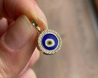 Pode incluir: Pingente dourado com um design de olho grego azul e branco. O pingente circular tem uma borda dourada com um padrão de chave grega. O olho tem um fundo azul com um centro branco e amarelo.