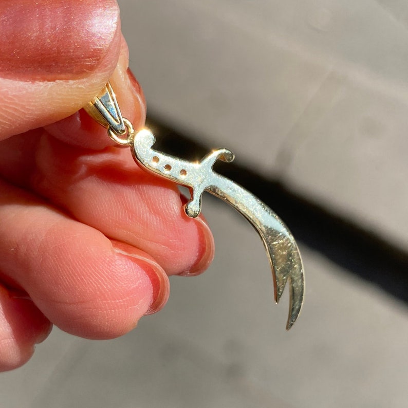 K&ouml;nnte beinhalten: Ein goldfarbener Anh&auml;nger in Form eines gebogenen Schwertes, vor unscharfem Hintergrund. Der Anh&auml;nger hat oben eine kleine Schlaufe f&uuml;r eine Kette und drei kleine L&ouml;cher entlang der Klinge.