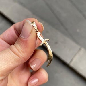 K&ouml;nnte beinhalten: Ein goldfarbener Anh&auml;nger in Form eines gebogenen Schwertes, gehalten von einer Hand. Der Anh&auml;nger hat oben eine Schlaufe f&uuml;r eine Kette und drei kleine Punkte in der N&auml;he des Griffs. Die Klinge des Schwertes ist gebogen und hat eine Spitze.