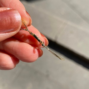 K&ouml;nnte beinhalten: Nahaufnahme eines goldfarbenen Ohrrings. Der Ohrring hat ein d&uuml;nnes, l&auml;ngliches Design mit einem kleinen dekorativen Element in der N&auml;he der Oberseite. Der Ohrring wird zwischen den Fingern gehalten, und der Hintergrund ist unscharf.