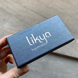 May include: A dark blue textured jewelry box with the word "likya" in silver lettering. The box also has the text "kuyumculuk" in silver lettering. The box is rectangular and has a slightly reflective surface.