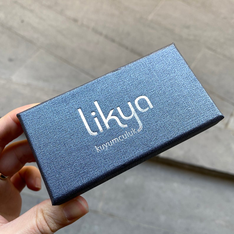 May include: A dark blue textured jewelry box with the word "likya" in silver lettering. The box also has the text "kuyumculuk" in silver lettering. The box is rectangular and has a slightly reflective surface.