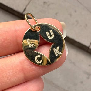 May include: Gold-colored pendant with the word "LUCK" spelled out in capital letters. The pendant has a circular design with the letters arranged around the center. The pendant is held by a person.
