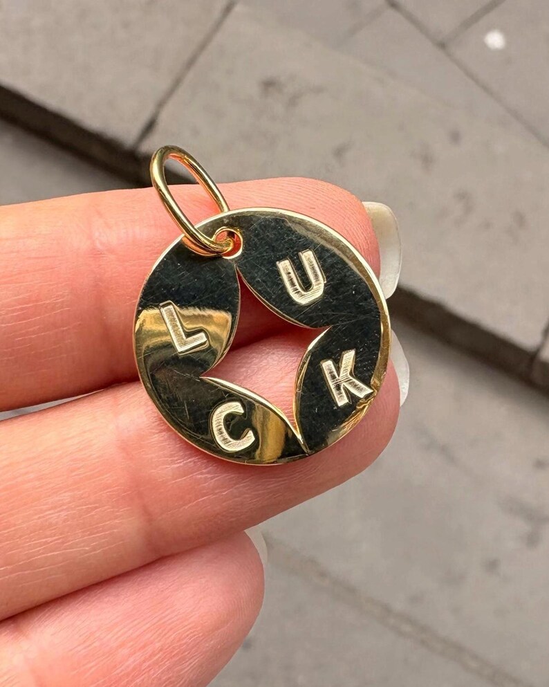 May include: Gold-colored pendant with the word "LUCK" spelled out in capital letters. The pendant has a circular design with the letters arranged around the center. The pendant is held by a person.