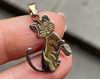 May include: A gold-toned cat pendant with a detailed outline. The pendant features a sitting cat with a curved tail and a loop for a chain. The cat's face has detailed eyes and whiskers. The pendant is held by a person's fingers.
