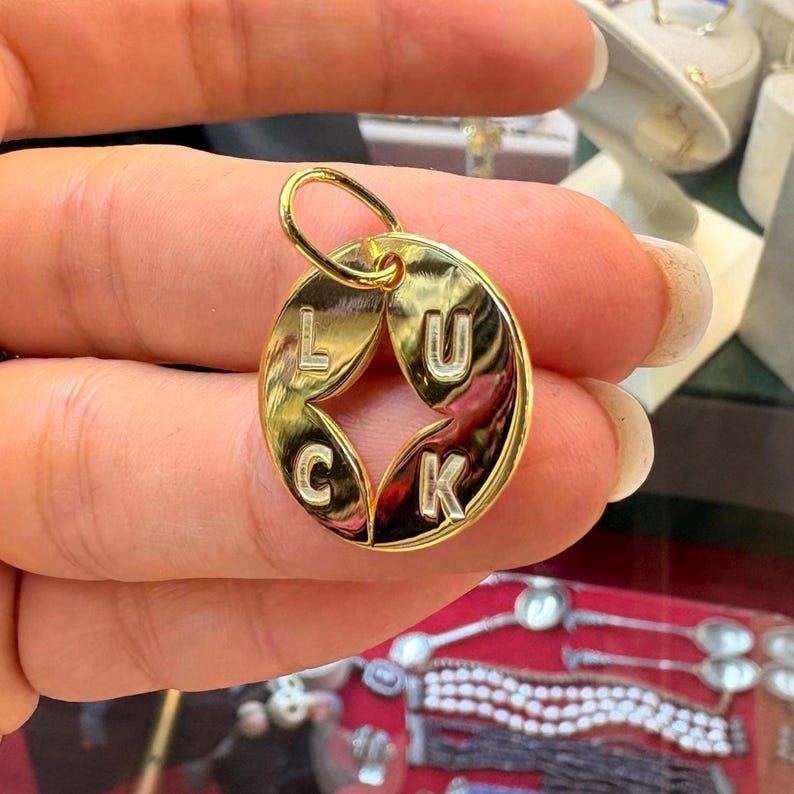 May include: A gold-colored pendant with the word "LUCK" split across two connected circular shapes. The letters are in a simple, sans-serif font. The pendant has a small loop at the top for a chain.