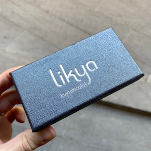 K&ouml;nnte beinhalten: Eine dunkelblaue, strukturierte Schmuckschatulle mit dem Wort "likya" in silbernen Buchstaben auf der Oberseite. Das Wort "kuyumculuk" ist unter "likya" aufgedruckt. Die Schachtel wird von einer Person in der Hand gehalten.