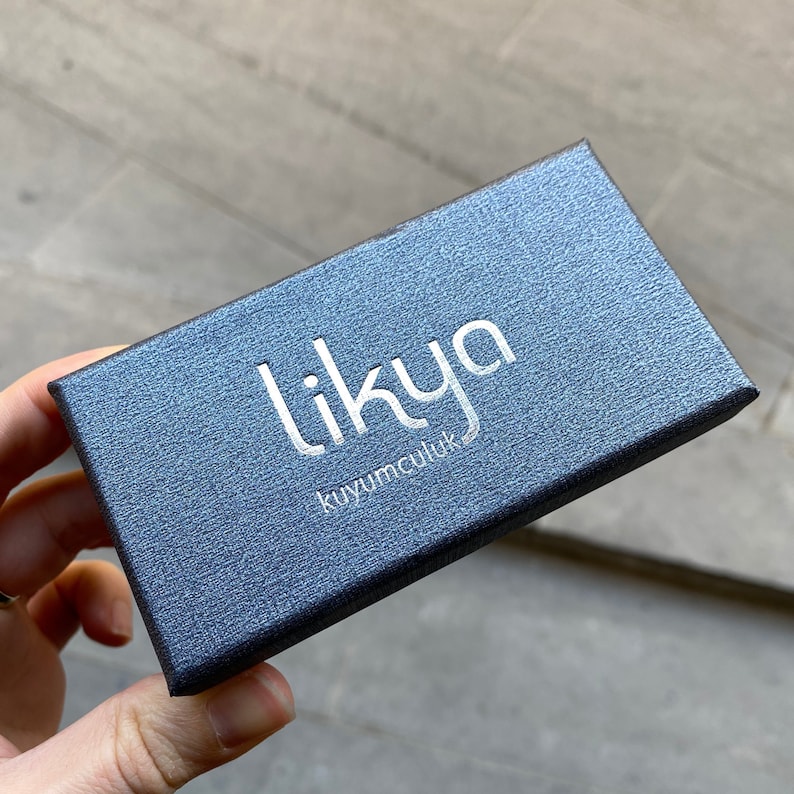 K&ouml;nnte beinhalten: Eine dunkelblaue, strukturierte Schmuckschatulle mit dem Wort "likya" in silbernen Buchstaben auf der Oberseite. Das Wort "kuyumculuk" ist unter "likya" aufgedruckt. Die Schachtel wird von einer Person in der Hand gehalten.
